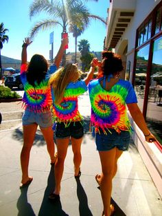 three girls in tie dye shirts are walking down the street and one girl is holding her hand up