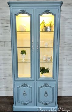 a blue china cabinet with glass doors and flowers in the top shelf, sitting on a wooden floor