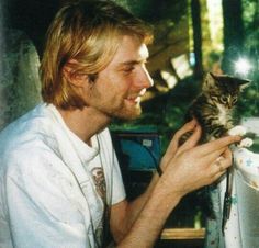 a man holding a kitten in his right hand and looking at the monitor screen while smiling