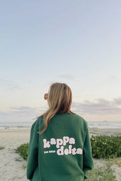 a woman standing on the beach wearing a green sweatshirt that says kappa dola