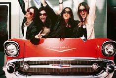 three women in sunglasses are sitting on the hood of an old red car and waving