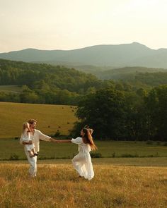 a man and woman holding hands while walking through a field with mountains in the background