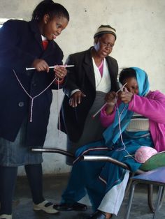 two women are knitting while another woman is sitting in a chair with her hand on the string