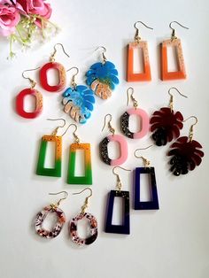 six pairs of earrings with different shapes and colors are shown on a white surface next to pink flowers