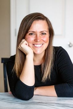 a smiling woman sitting at a table with her hand on her chin and looking into the camera