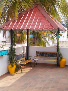 two benches sitting under a red roof next to palm trees and potted plants on the side of a building