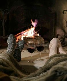 two glasses of wine are sitting in front of an open fire with feet propped up