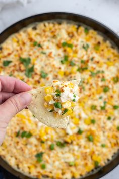 a hand holding a tortilla chip over a skillet filled with corn and cheese