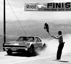 a man holding a flag next to a car on top of a dirt road with mountains in the background