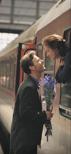 a man and woman standing next to a train with flowers in their hands, looking out the window