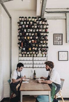 two men sitting at a table in front of a wall with wine bottles on it