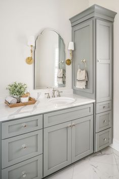 a bathroom with gray cabinets and white marble counter tops, gold accents on the mirror