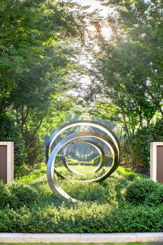 an abstract sculpture in the middle of a park surrounded by bushes and trees with sunlight shining through