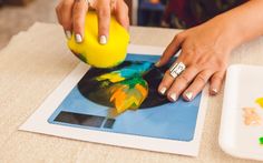 a woman is painting with yellow and blue colors on the table next to a white plate