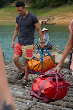 two men carrying luggage across a wooden bridge next to a body of water with boats in the background