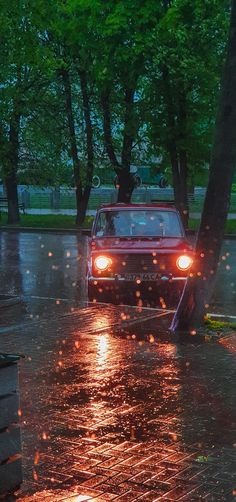 a red car driving down a rain soaked street next to trees with lights on it