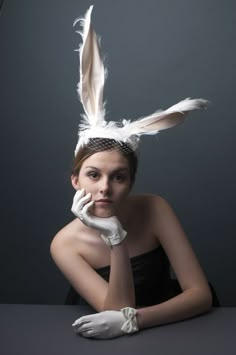 a woman with white gloves and feathers on her head is sitting at a table in front of a gray background