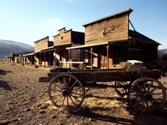 an old wooden wagon sitting in the middle of a dirt field next to some buildings