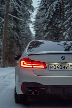 the back end of a white bmw car driving down a snow covered road with trees in the background