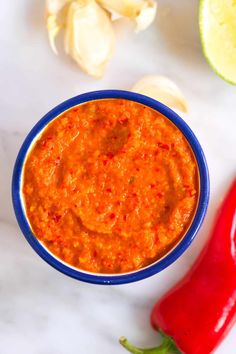 a blue bowl filled with red sauce next to an orange pepper, garlic and lime