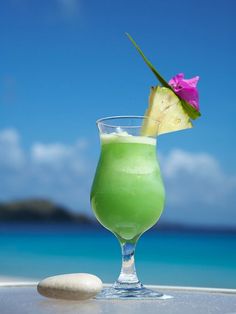 a green drink sitting on top of a white table next to the ocean with a pink flower in it