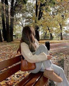 a woman sitting on top of a wooden bench in the fall leaves with her legs crossed