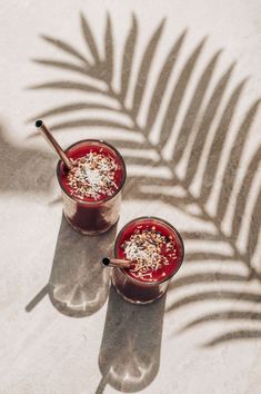 two glasses filled with red liquid and topped with sprinkles next to a palm leaf