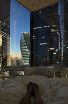 a person laying in bed with their feet on the pillow looking out at skyscrapers