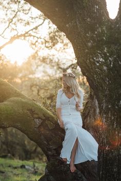 a woman in a white dress is sitting on a tree branch and posing for the camera