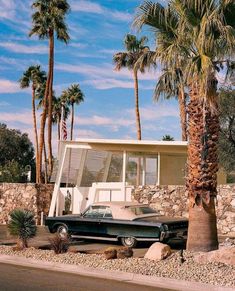 an old car parked in front of a house with palm trees and rocks around it