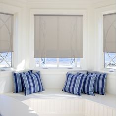 a window seat with pillows on it in front of two windows that have roman shades over them