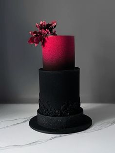 a three tiered black cake with pink frosting and flowers on top, sitting on a marble table