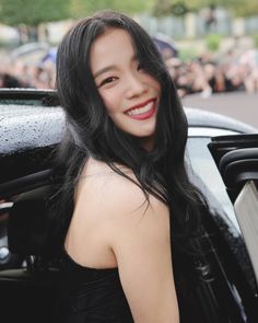 a woman with long black hair standing next to a car and smiling at the camera