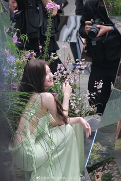 a woman in a green dress sitting on a bench next to flowers and people taking pictures