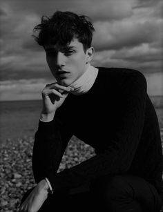 a young man sitting on the beach with his hand under his chin and looking at the camera