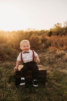 a little boy sitting on top of a wooden crate in the grass wearing a bow tie and suspenders