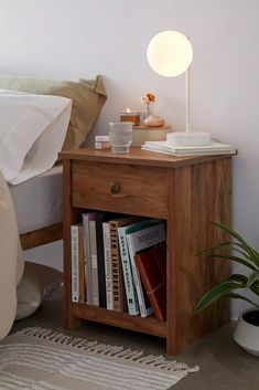 a nightstand with books on it next to a bed and a potted plant in the corner