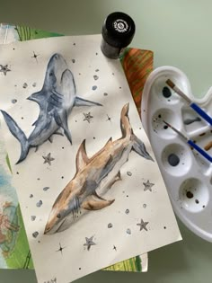 two shark paintings on paper next to paintbrushes and watercolor paints with brushes