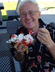 an older man sitting at a table with food in his hand and smiling for the camera