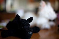 two small stuffed animals sitting on top of a wooden table next to each other with blurry cats in the background