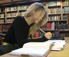 a woman sitting at a table in front of a book shelf holding a pen and writing