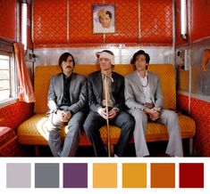 three men in suits sitting on a bus with orange and red walls, one man is looking at the camera