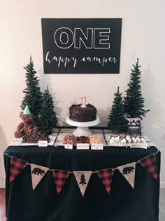 a black table topped with a cake covered in frosting and pine trees next to a sign that says one happy camper