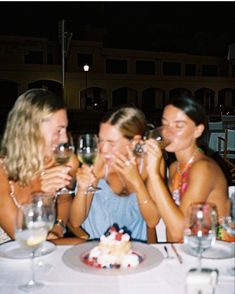 three women sitting at a table with wine glasses in front of their faces and smiling