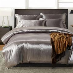 a bed with silver sheets and pillows in a room next to two windows, one has a brown throw blanket on it