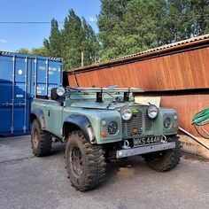 an army green jeep parked in front of a blue dumpster next to a wooden wall