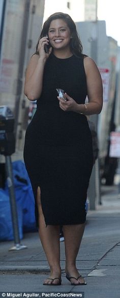 a woman in a black dress talking on a cell phone while walking down the street