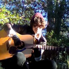 a young man is playing an acoustic guitar in front of some trees and bushes,