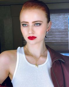 a woman with red lipstick wearing a white tank top and brown leather jacket is looking at the camera