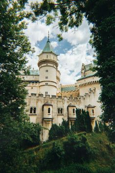 the castle is built on top of a hill surrounded by trees and bushes, under a cloudy blue sky with white clouds